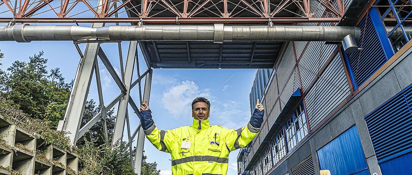 Mann mit neongelber Schutzjacke steht vorm Biomasse Heizkraftwerk und zeigt nach oben