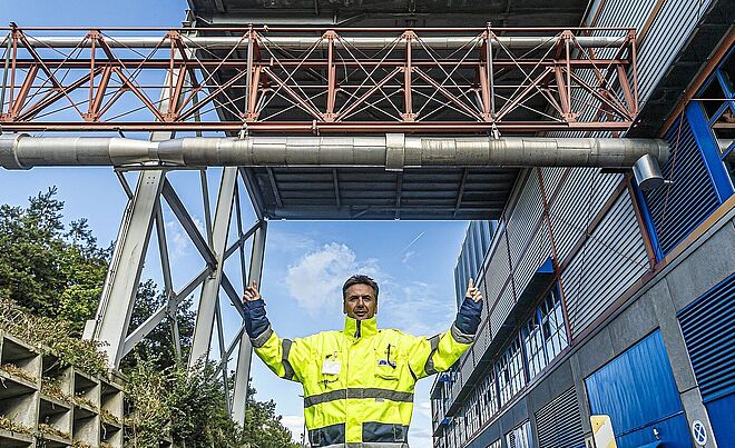 Mann mit neongelber Schutzjacke steht vorm Biomasse Heizkraftwerk und zeigt nach oben