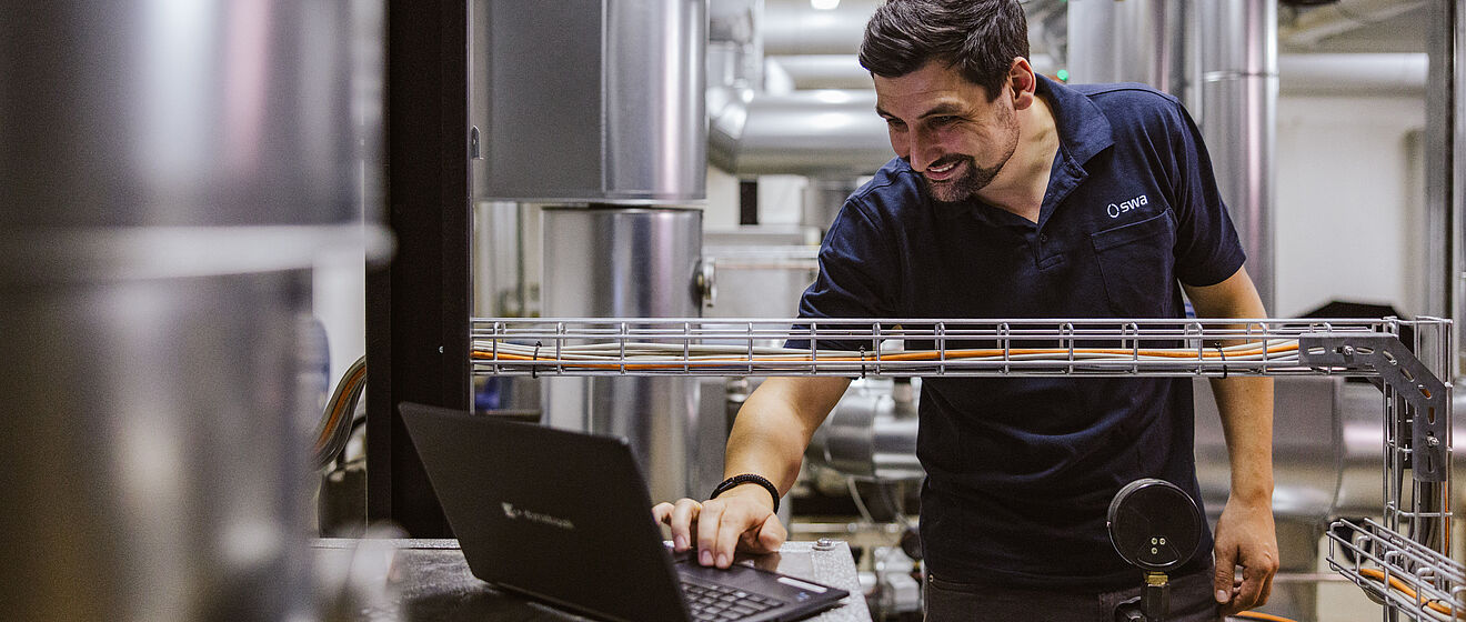 Mann tippt an einem Laptop und ist umgeben von Gebäudetechnik