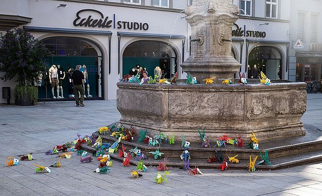 Eine öffentliche Installation aus vielen bunten, gefalteten Papierfiguren steht rund um einen Brunnen vor einem Geschäft namens "Eckerle Studio" in der Augsburger Innenstadt.