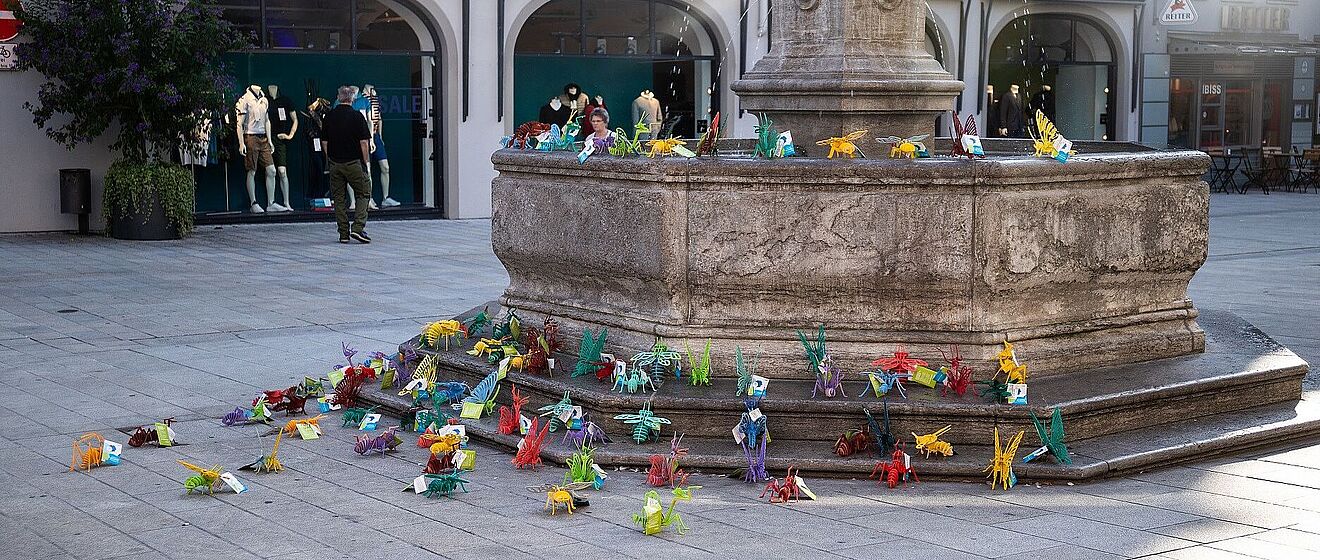 Eine öffentliche Installation aus vielen bunten, gefalteten Papierfiguren steht rund um einen Brunnen vor einem Geschäft namens "Eckerle Studio" in der Augsburger Innenstadt.