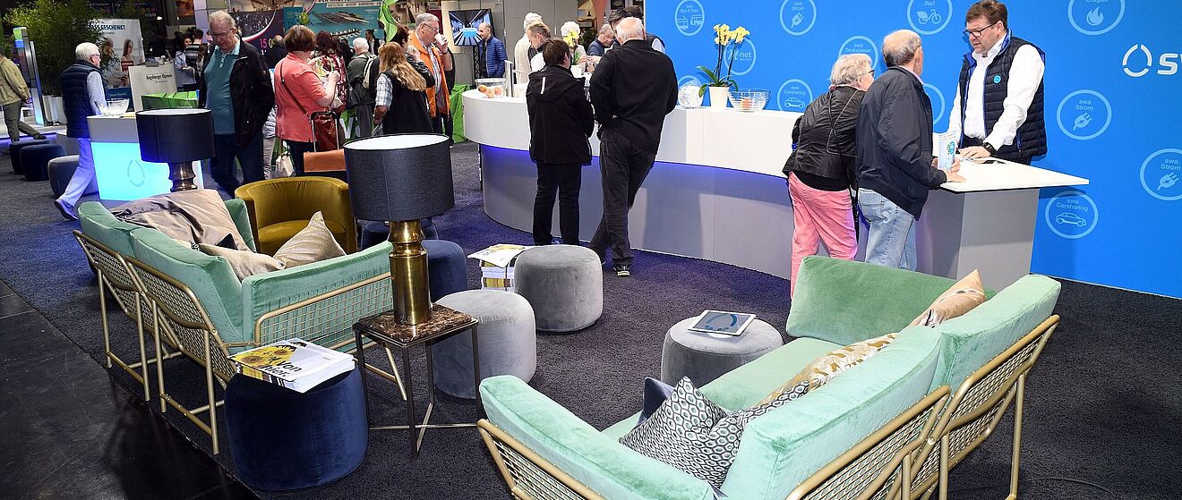 Im Vordergrund ist eine Reihe an Sesseln und anderen Sitzgelegenheiten. Im Hintergrund befindet sich ein Stand von den Stadtwerken Augsburg. Dort stehen mehrere Besucher der afa und unterhalten sich mit Personen in swa-Jacken.