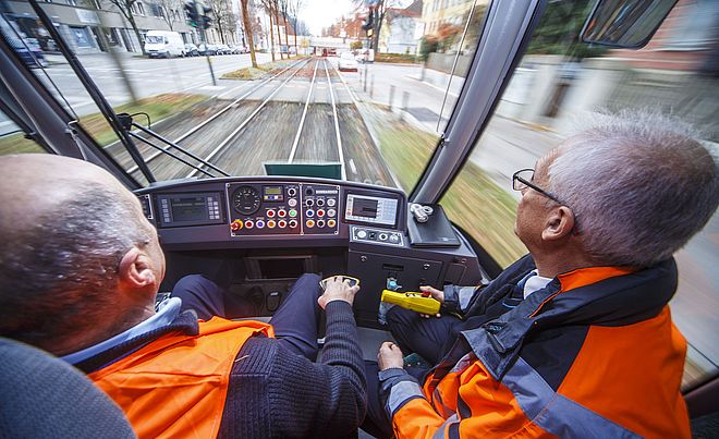 Blick aus der Fahrerkabine einer Straßenbahn. Im Vordergrund sitzen zwei Männer in Warnwesten, die die Steuerelemente bedienen.