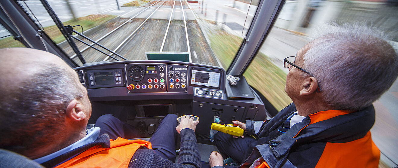 Blick aus der Fahrerkabine einer Straßenbahn. Im Vordergrund sitzen zwei Männer in Warnwesten, die die Steuerelemente bedienen.