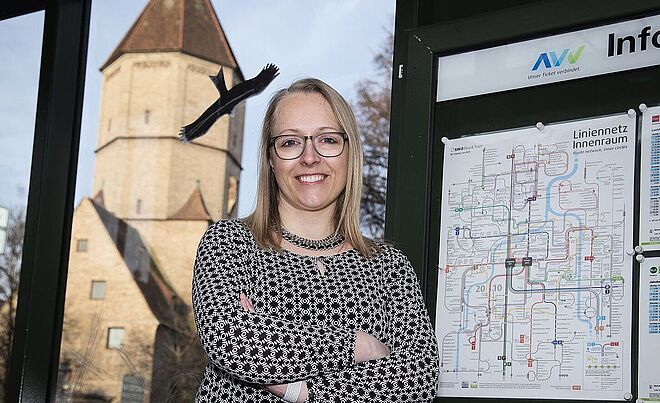 Eine Frau mit Brille und einem schwarz-weißen, gemusterten Oberteil steht in Augsburg neben einer Informationstafel mit dem Logo des Augsburger Verkehrsverbunds (AW) und dem Text "Liniennetz Innenraum". Hinter ihr ist durch das Fenster das historische Jakobertor sichtbar.