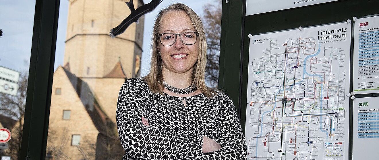 Eine Frau mit Brille und einem schwarz-weißen, gemusterten Oberteil steht in Augsburg neben einer Informationstafel mit dem Logo des Augsburger Verkehrsverbunds (AW) und dem Text "Liniennetz Innenraum". Hinter ihr ist durch das Fenster das historische Jakobertor sichtbar.