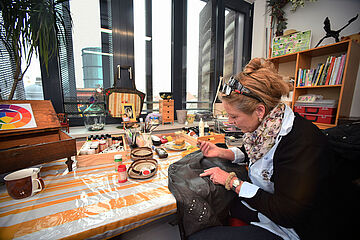 Frau sitzt an einem Tisch mit verschiedenen Farbutensilien und arbeitet an einer Lederjacke. Ein Klick auf das Bild führt zu einer größeren Ansicht.