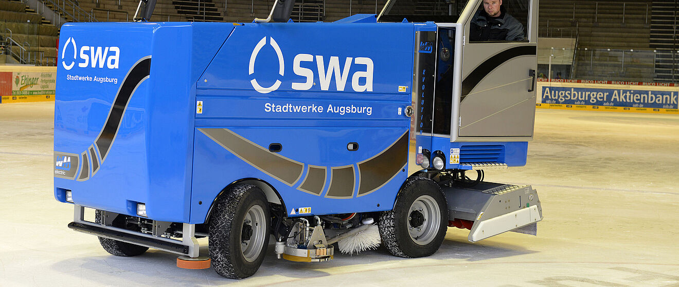 Eine blaue Eismaschine der Stadtwerke Augsburg steht im Vordergrund auf einer Eisfläche in einem Stadion. Ein Mann steuert das Fahrzeug, während im Hintergrund die leeren Ränge der Arena sichtbar sind.