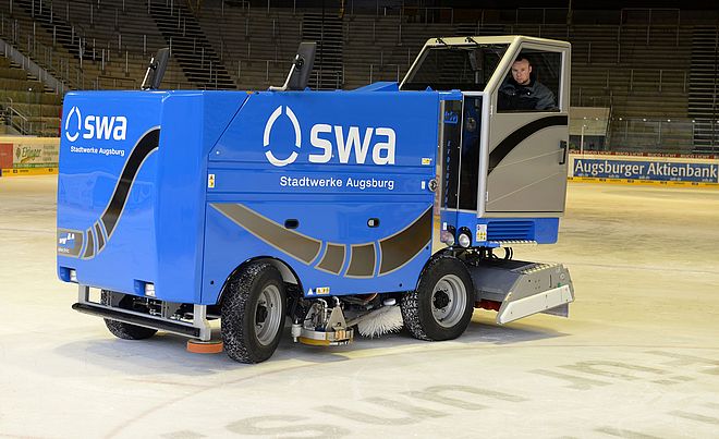 Eine blaue Eismaschine der Stadtwerke Augsburg steht im Vordergrund auf einer Eisfläche in einem Stadion. Ein Mann steuert das Fahrzeug, während im Hintergrund die leeren Ränge der Arena sichtbar sind.