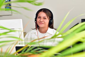 Eine junge Frau sitzt am Schreibtisch vor einem Computerbildschirm. Sie trägt ein Headset. Zudem sind Blätter einer Büropflanze zu sehen.