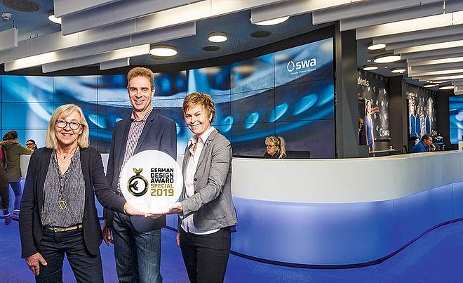 Zwei Frauen und ein Mann präsentieren den German Design Award Special 2019. Im Hintergrund befindet sich das moderne swa Kundencenter mit beleuchteten Theken und Medienwand.