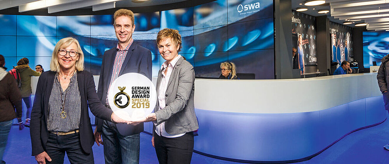 Zwei Frauen und ein Mann präsentieren den German Design Award Special 2019. Im Hintergrund befindet sich das moderne swa Kundencenter mit beleuchteten Theken und Medienwand.