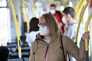 Frau steht mit Maske in einer Straßenbahn - Ein Klick auf das Bild führt zur vergrößerten Fotoansicht.