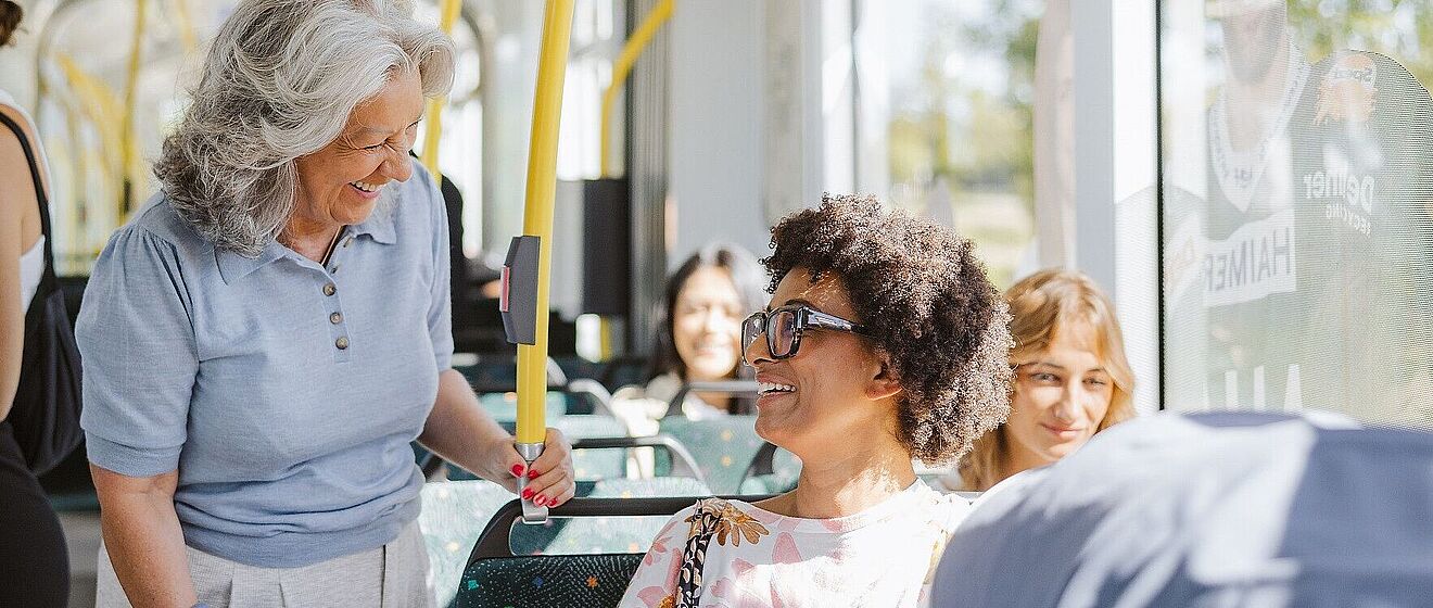 Eine grinsende Frau, die in einer Straßenbahn sitzt und von einer stehenden älteren Dame angelächelt wird.  Mit Link zum öffnen einen Pop-Ups mit Informationen zum Mobiil-Abo Premium