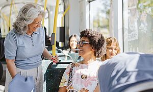 Eine grinsende Frau, die in einer Straßenbahn sitzt und von einer stehenden älteren Dame angelächelt wird.  Mit Link zum öffnen einen Pop-Ups mit Informationen zum Mobiil-Abo Premium