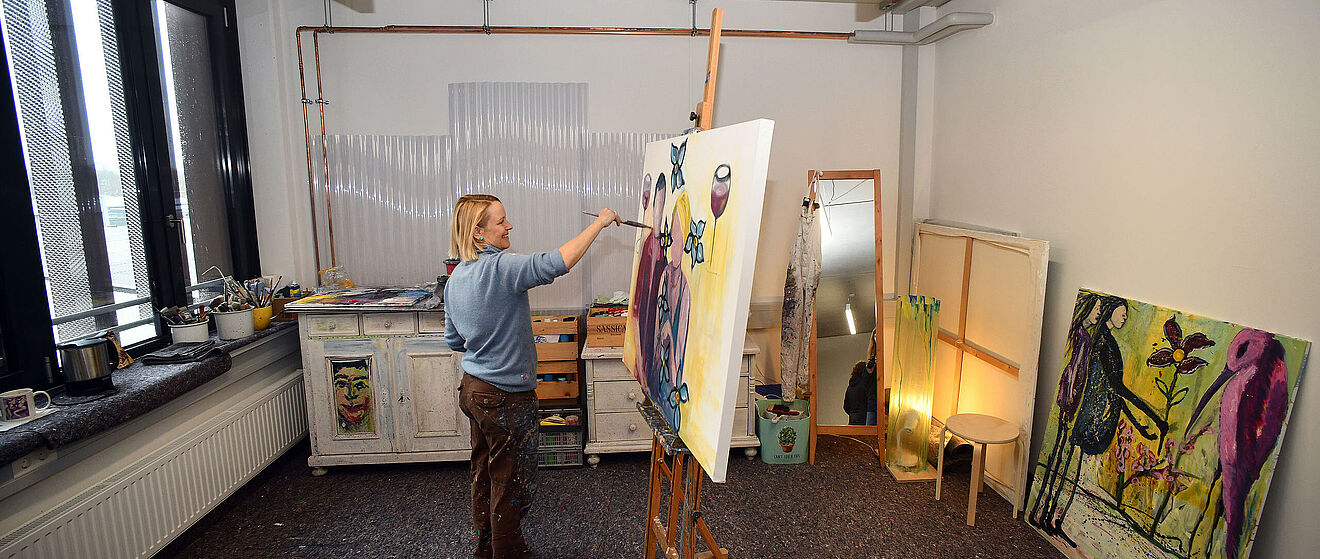 Frau steht in einem Raum mit Pinsel in der Hand vor einer Leinwand und malt ein Gemälde