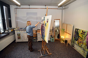 Frau steht in einem Raum mit Pinsel in der Hand vor einer Leinwand und malt ein Gemälde - Ein Klick auf das Bild führt zur vergrößerten Fotoansicht.