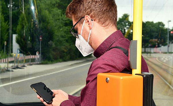 Mann sitzt mit Mundschutz in Straßenbahn und schaut auf sein Handy
