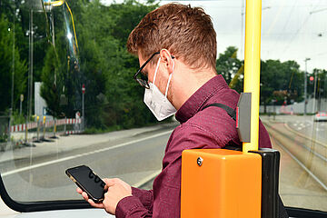 Mann sitzt mit Mundschutz in Straßenbahn und schaut auf sein Handy - Beim anklicken wird das Bild vergrößert.