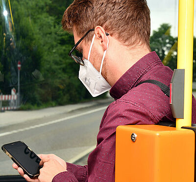 Mann sitzt mit Mundschutz in Straßenbahn und schaut auf sein Handy