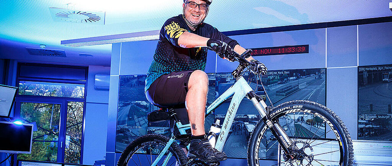 Ein lächelnder Mann sitzt in Fahrradausrüstung auf einem Mountainbike. Im Hintergrund befindet sich eine Wand mit einer Reihe von Bildschirmen.