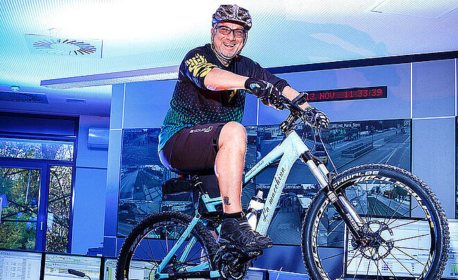Ein lächelnder Mann sitzt in Fahrradausrüstung auf einem Mountainbike. Im Hintergrund befindet sich eine Wand mit einer Reihe von Bildschirmen.