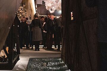 Ein Blickwinkel ins Foyer des Ofenhauses, in dem sich mehrere vornehm gekleidete Personen befinden. - Ein Klick auf das Bild führt zur vergrößerten Fotoansicht.