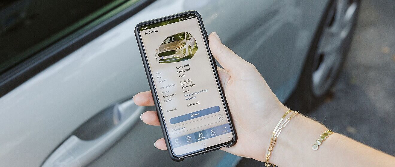 Hand hält Smartphone mit geöffneter Carsharing App. Im Hintergrund ist ein Auto.
