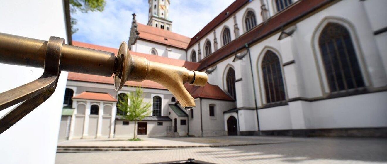 Trinkwasserbrunnen bei St. Ulrich in Augsburg. Im Hintergrund ist eine Kirche
