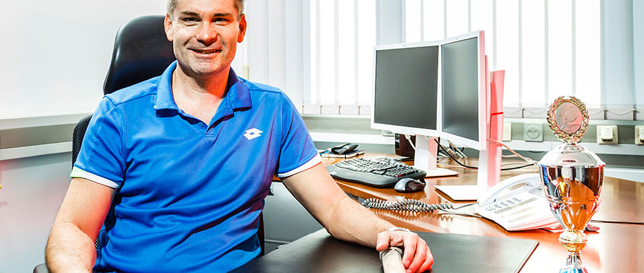 Ein Mann schaut lächelnd in die Kamera. Er sitzt an seinem Bürotisch und hält einen Tennisschläger in der Hand. Auf dem Bürotisch befindet sich eine Tennistrophäe.