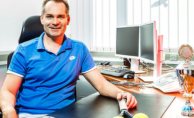 Ein Mann schaut lächelnd in die Kamera. Er sitzt an seinem Bürotisch und hält einen Tennisschläger in der Hand. Auf dem Bürotisch befindet sich eine Tennistrophäe.