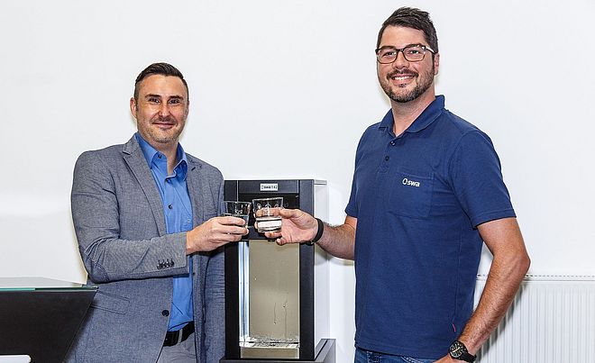 Zwei Männer stehen neben einer Tafelwasseranlage der swa und stoßen mit zwei Wassergläsern an. Der rechte Mann trägt Kleidung von den Stadtwerken Augsburg.