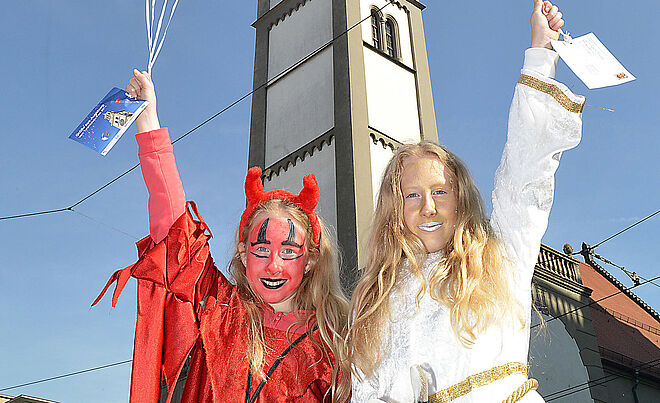 Ein Mädchen im Teufelskostüm und ein Mädchen mit Goldschminke im Gesicht heben jeweils eine Hand und stehen vor dem Perlachturm