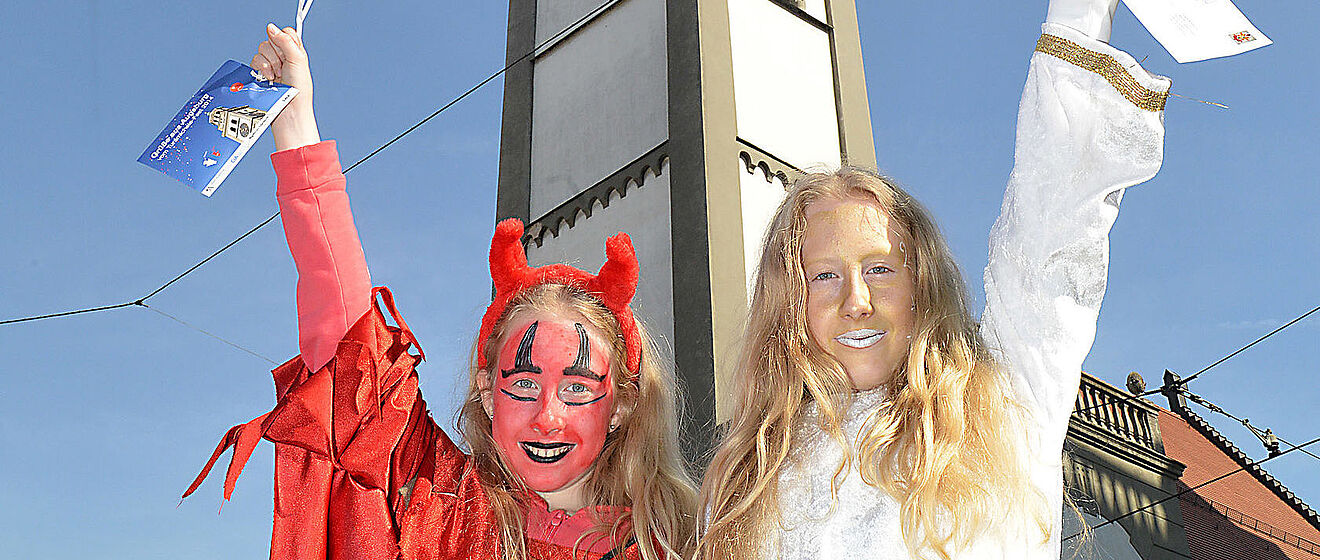 Ein Mädchen im Teufelskostüm und ein Mädchen mit Goldschminke im Gesicht heben jeweils eine Hand und stehen vor dem Perlachturm