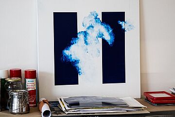 Im Vordergrund auf einer Metallablage liegen Künstlerbedarf und Werkzeuge, darunter zwei rote Sprühdosen, eine offene Packung Klebeband und ein rot-weißer Werkzeugkasten. In der Bildmitte steht ein abstraktes Gemälde auf einer Staffelei, das blaue, wolkenartige Farbwolken auf weißem Hintergrund zwischen zwei vertikalen schwarzen Balken zeigt. Ein Klick auf das Bild führt zu einer größeren Ansicht.