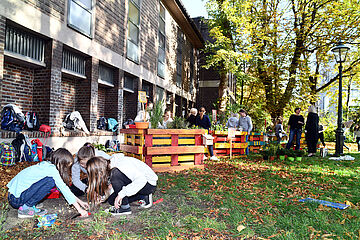 Vier Mädchen beugen sich im Vordergrund über den Boden, um etwas einzupflanzen, während im Hintergrund Erwachsene zu sehen sind. Ein Klick auf das Bild führt zu einer größeren Ansicht.