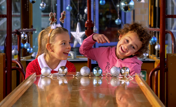 Zwei Kinder sitzen in der weihnachtlich geschmückten Christkindltram der Stadtwerke Augsburg an einem Holztisch, der mit glänzenden silbernen und blauen Christbaumkugeln dekoriert ist.