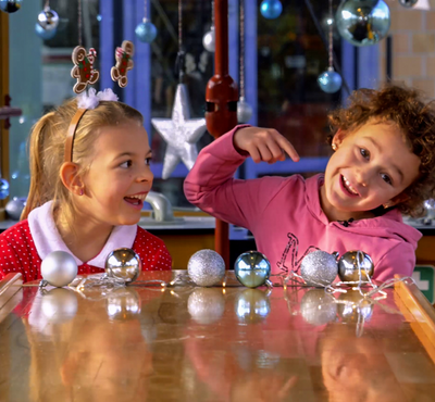 Zwei Kinder sitzen in der weihnachtlich geschmückten Christkindltram der Stadtwerke Augsburg an einem Holztisch, der mit glänzenden silbernen und blauen Christbaumkugeln dekoriert ist.