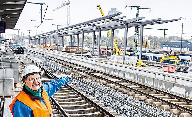 Eine Frau mit Schutzhelm zeigt lächelnd auf den sich im Hintergrund befindlichen Bahnsteig F mit zwei Gleisen. Im Hintergrund befindet sich eine Baustelle des neuen Augsburger Hauptbahnhofs.   