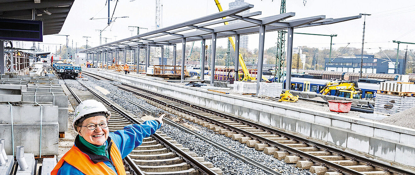 Eine Frau mit Schutzhelm zeigt lächelnd auf den sich im Hintergrund befindlichen Bahnsteig F mit zwei Gleisen. Im Hintergrund befindet sich eine Baustelle des neuen Augsburger Hauptbahnhofs.