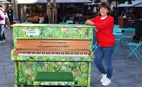 Bemaltes Klavier, in Grün Tönen. Frau lehnt sich an das Klavier an. 