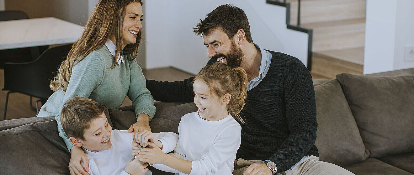 Eine lachende Frau hinter einem Sofa und ein lachender Mann und zwei lachende Kinder auf einem Sofa