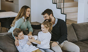 Eine lachende Frau hinter einem Sofa und ein lachender Mann und zwei lachende Kinder auf einem Sofa