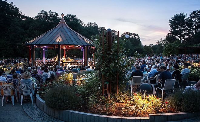 Mann spielt Musik in einem Pavillon. Um ihn herum sitzt ein großes Publikum. 