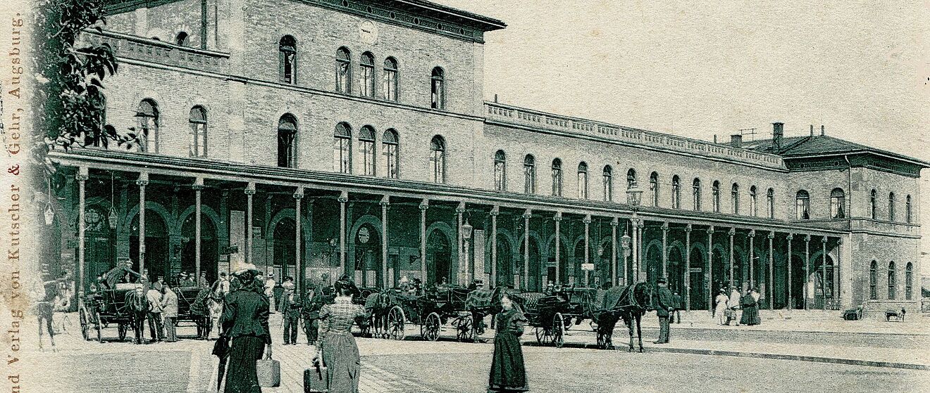 Das historische Bild zeigt das alte, repräsentative Empfangsgebäude des Augsburger Hauptbahnhofs. Auf dem davorliegenden, gepflasterten Platz, der um das Jahr 1900 aufgenommen wurde, sind mehrere Pferdekutschen und Menschen in zeitgenössischer Kleidung zu sehen, was das Alter der Aufnahme unterstreicht.