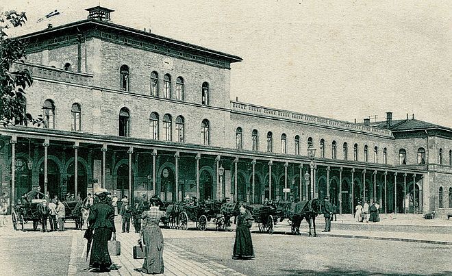 Das historische Bild zeigt das alte, repräsentative Empfangsgebäude des Augsburger Hauptbahnhofs. Auf dem davorliegenden, gepflasterten Platz, der um das Jahr 1900 aufgenommen wurde, sind mehrere Pferdekutschen und Menschen in zeitgenössischer Kleidung zu sehen, was das Alter der Aufnahme unterstreicht.