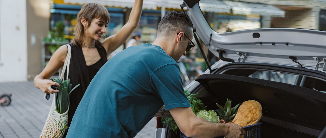 Mann mit Einkaufskorb belädt den Kofferaum von Carsharing Auto während eine Frau die Tür hält
