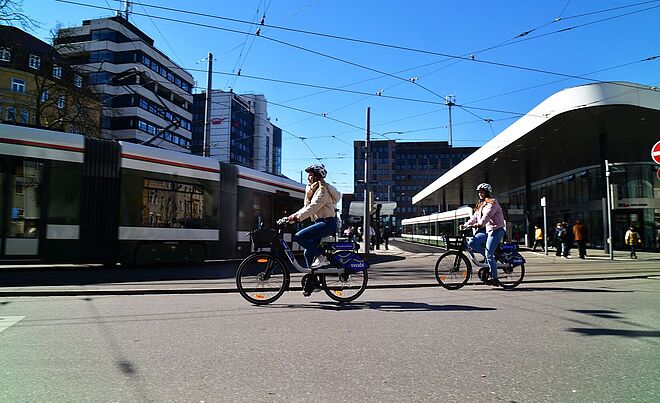 Zwei Frauen mit Helm fahren jeweils auf einem swabi am Königsplatz. Im Hintergrund fahren Straßenbahnen in die Haltestelle.