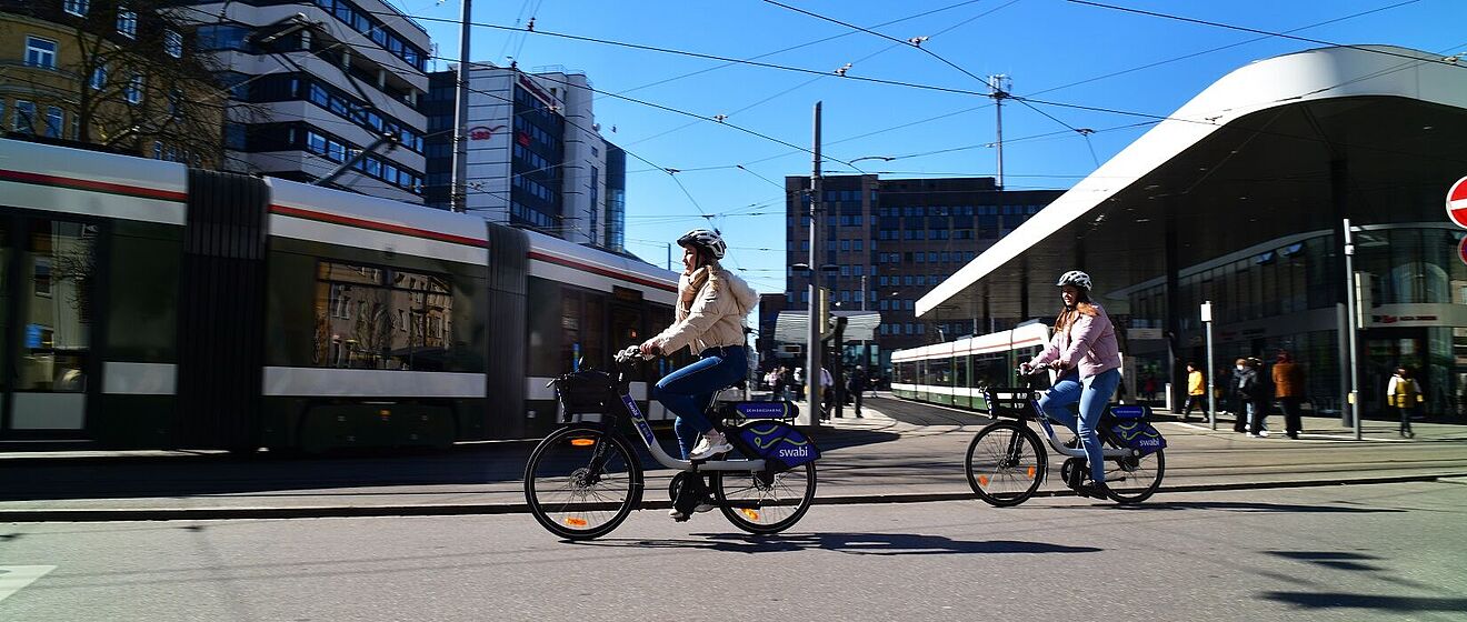 Zwei Frauen mit Helm fahren jeweils auf einem swabi am Königsplatz. Im Hintergrund fahren Straßenbahnen in die Haltestelle.