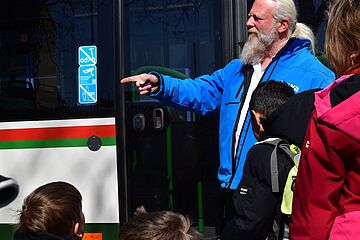 Mann mit Bart und swa Jacke zeigt auf ein Kind beim Bussicherheitstraining. Im Hintergrund ist ein Bus